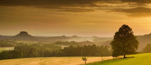 Blick über grünes Tal mit verstreuten Häusern und Hügeln im Hintergrund unter orangenem Himmel.