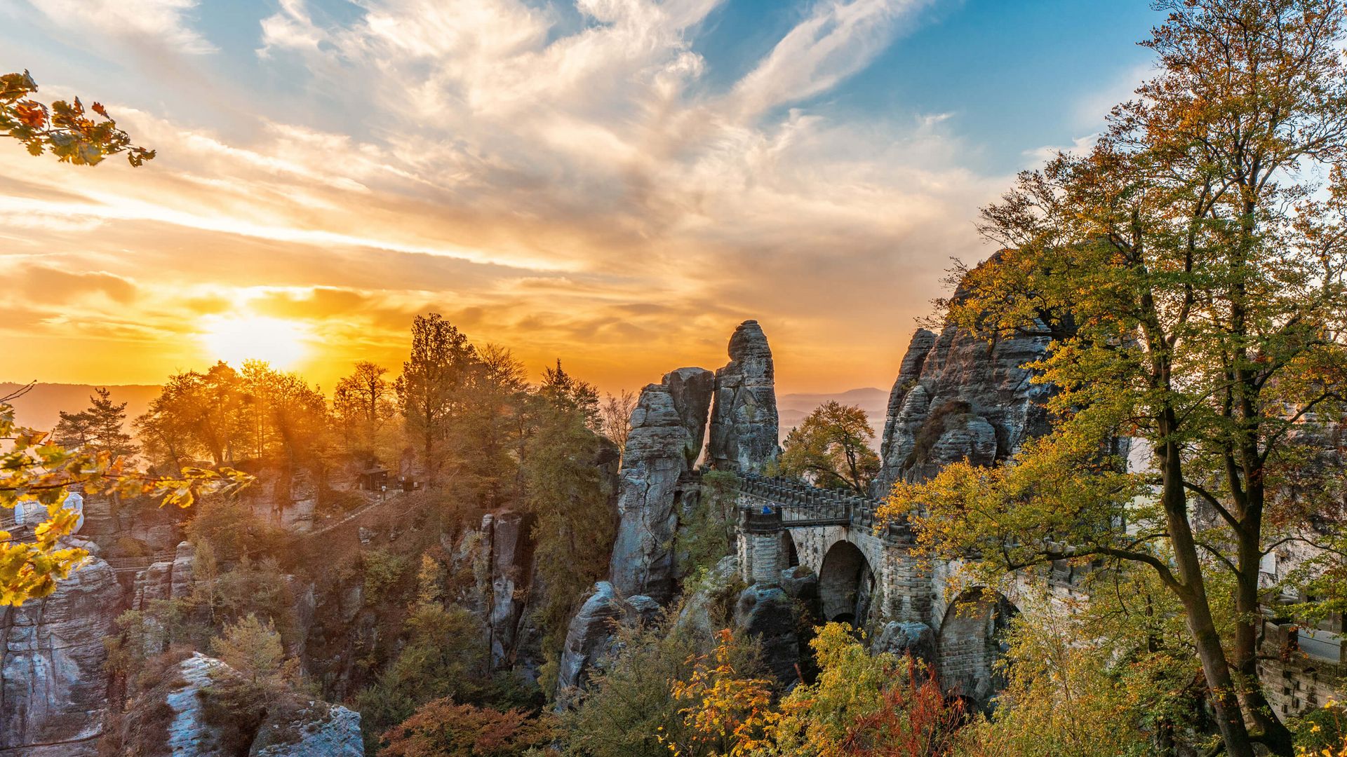 Sonnenuntergang hinter der Basteibrücke, umgeben von nebligem Wald und Sandsteinfelsen.