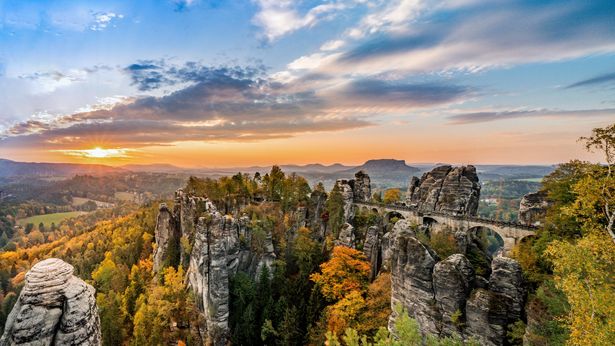 Felsformationen der Bastei mit Brücke, grüne Wälder, orange, blauer Himmel mit Sonne.