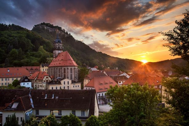 Sonnenuntergang über Festung Königstein, Kirche und Häuser im Vordergrund, orange-rosa Himmel.